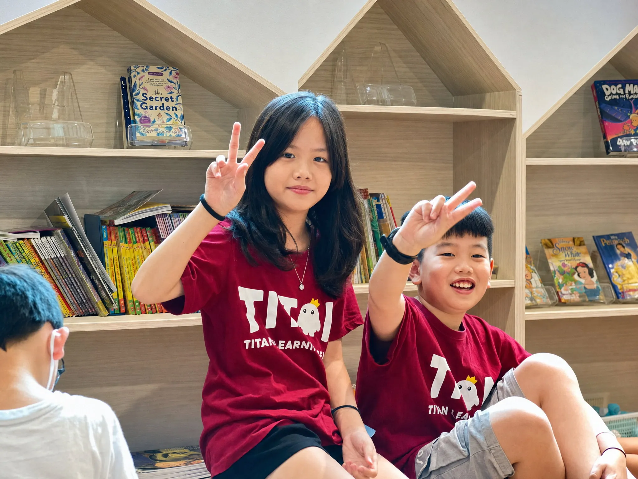 Two TITAN students sitting in front of a house-shaped bookcase of English storybooks, both making a peace sign at the camera