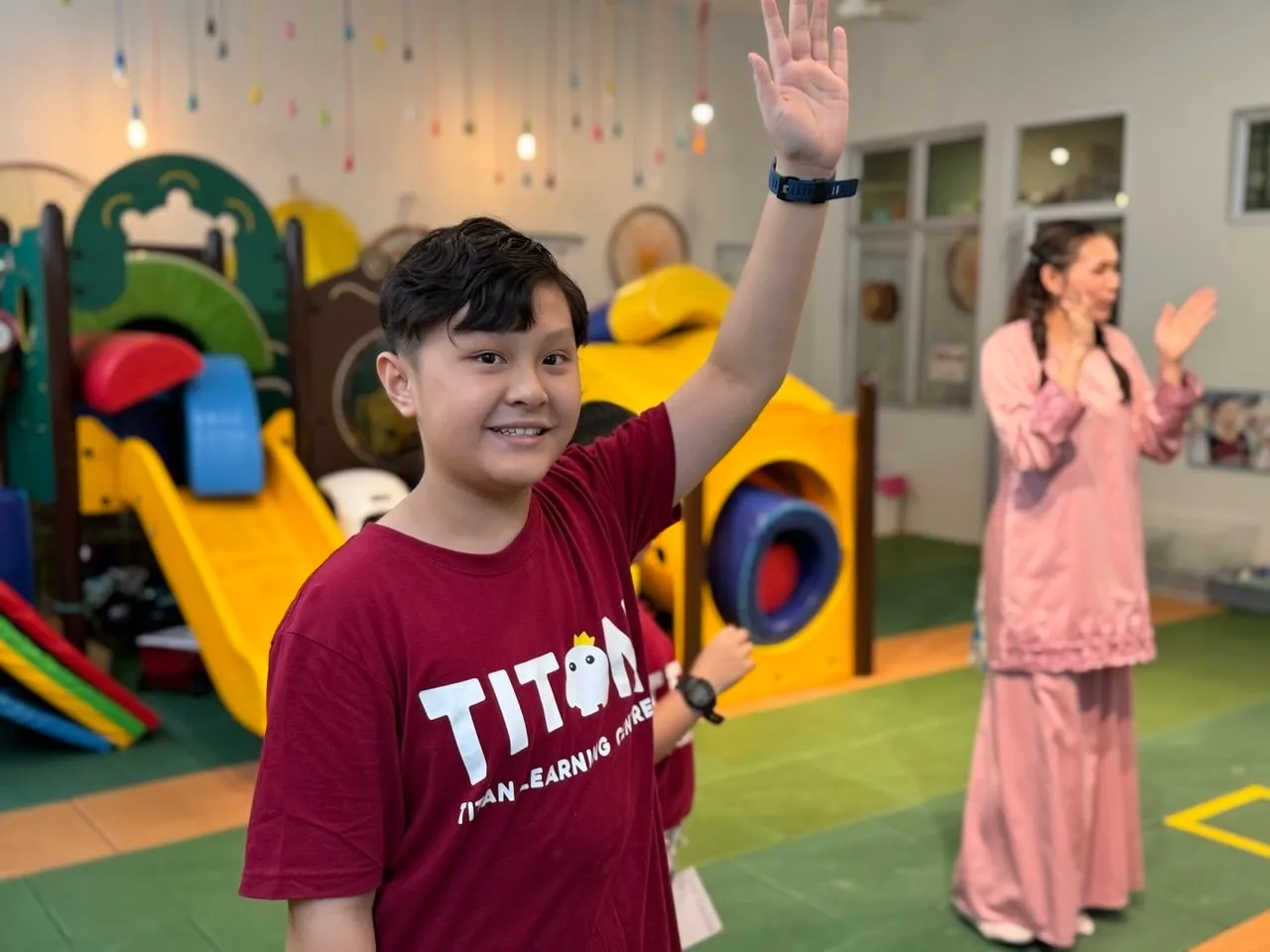 A TITAN student in a TITAN T-shirt with his hand raised, smiling, in the centre's activity room