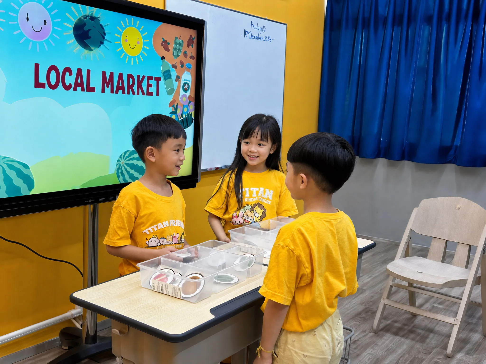 Three TITAN students in yellow shirts talking to each other across a table of props, during a Local Market role-play activity