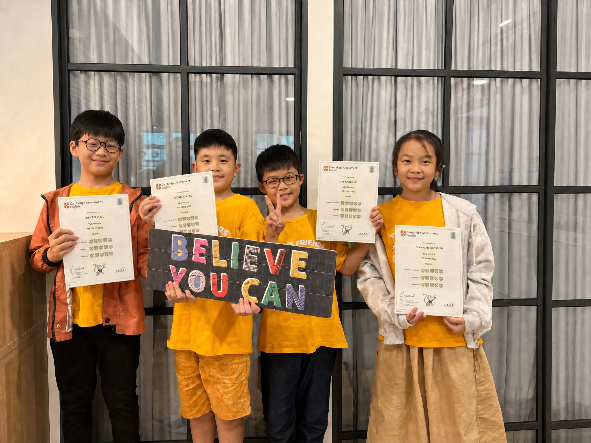 Four TITAN students holding their Cambridge Assessment English Young Learners' certificates, beside a board reading Believe You Can